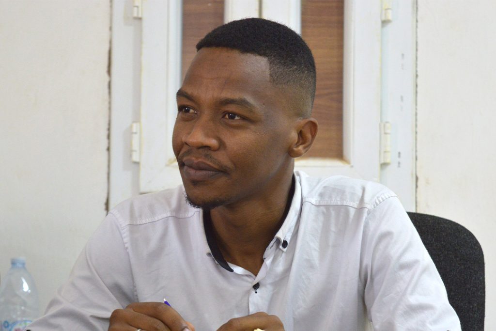 Asalik sitter på en kontorsstol och tittar snett framåt, förbi kameran. Han har en vit skjorta på sig.