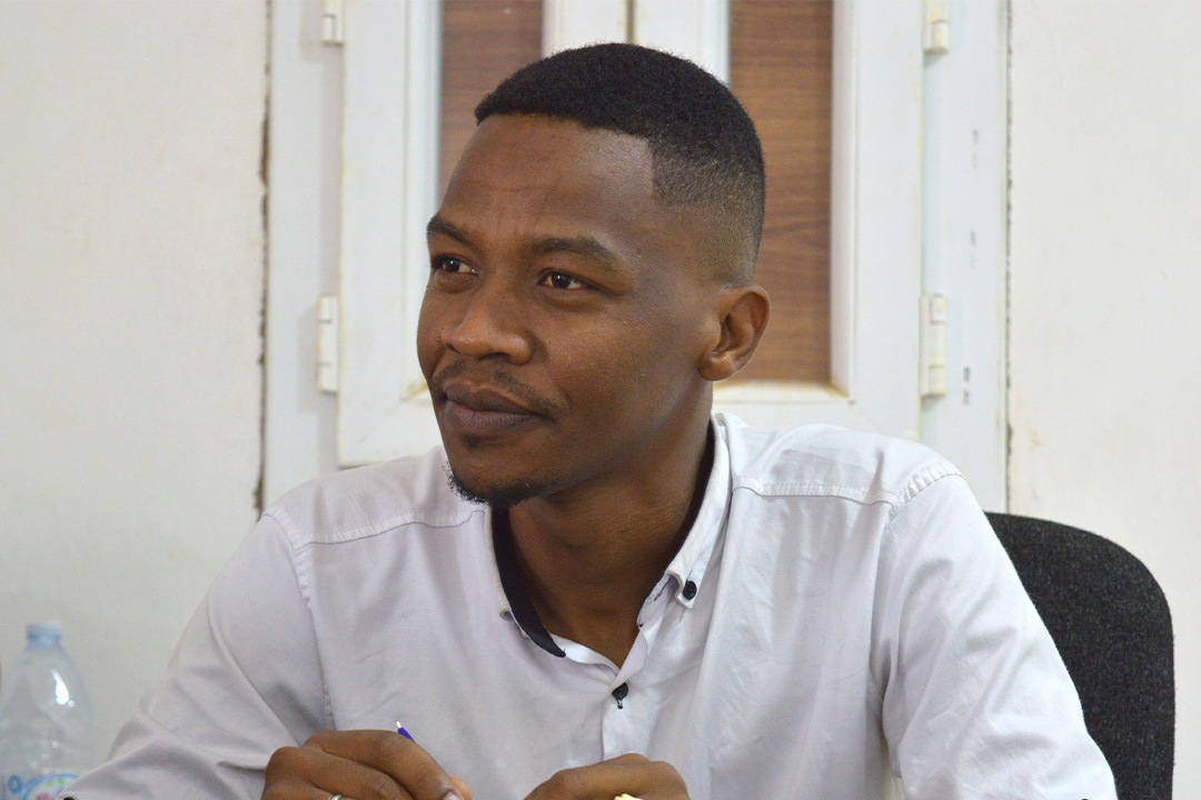 Asalik sitter på en kontorsstol och tittar snett framåt, förbi kameran. Han har en vit skjorta på sig.