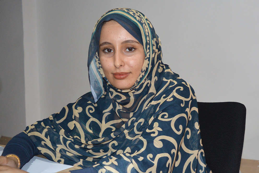 Asma sitter på en kontorsstol och tittar rakt in i kameran. Hon bär det traditionella västsahariska klädesplagget melfa.